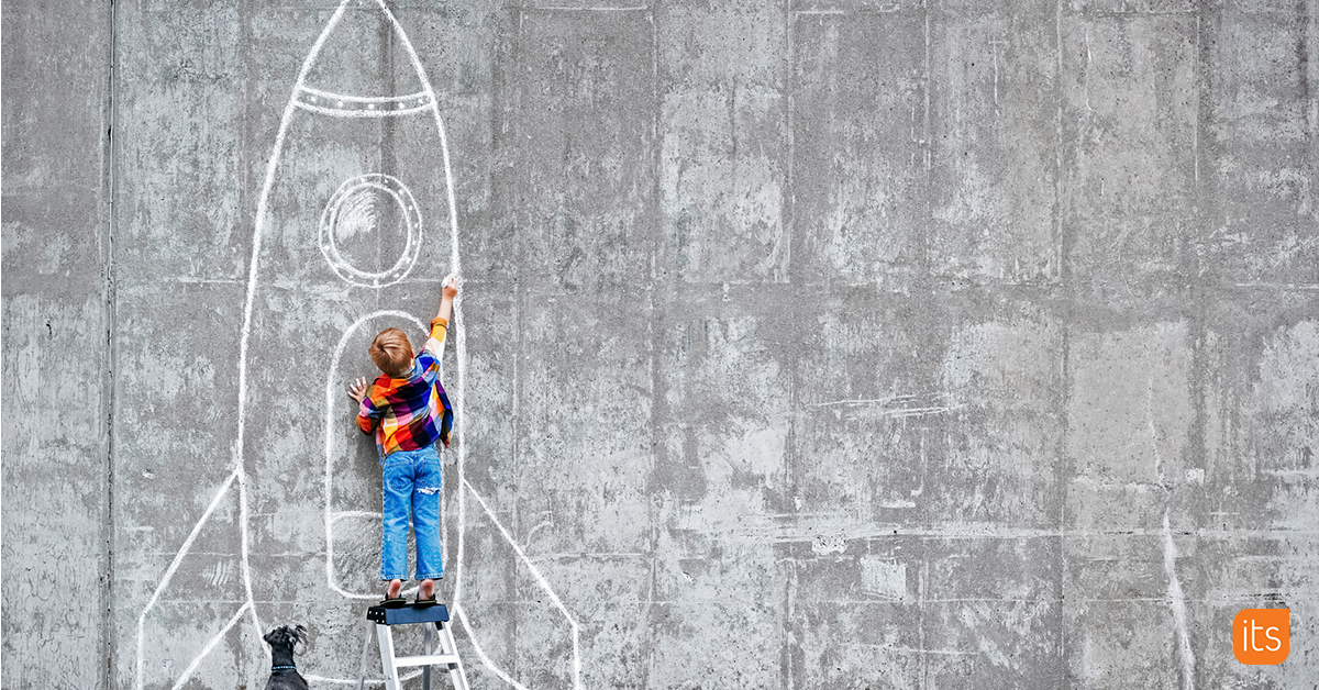 Photo of a student standing infront of a concrete wall with a chalk stick in hand, drawing a rocket ship.